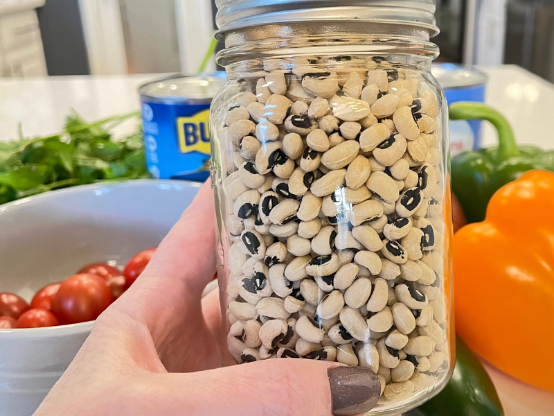 dry black-eyed peas in a glass jar with ingredients for cowboy caviar behind the peas.