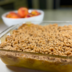 peach crisp with fresh peaches baked in square glass pan with a white bowl of whole peaches behind the crisp.