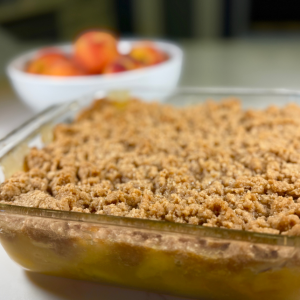 peach crisp with fresh peaches baked in square glass pan with a white bowl of whole peaches behind the crisp.