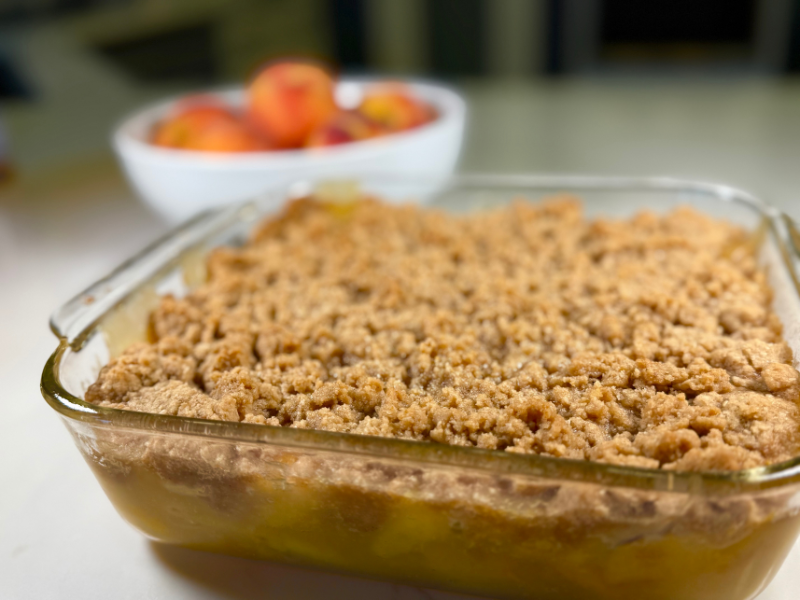 peach crisp with fresh peaches baked in square glass pan with a white bowl of whole peaches behind the crisp.