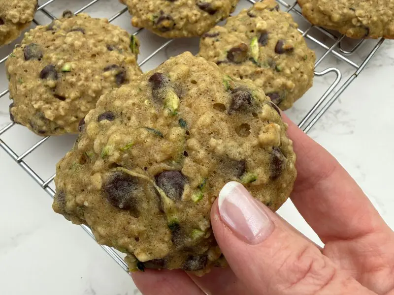 image of a hand holding a zucchini oatmeal chocolate chip cookie above a cooling rack of more cookies